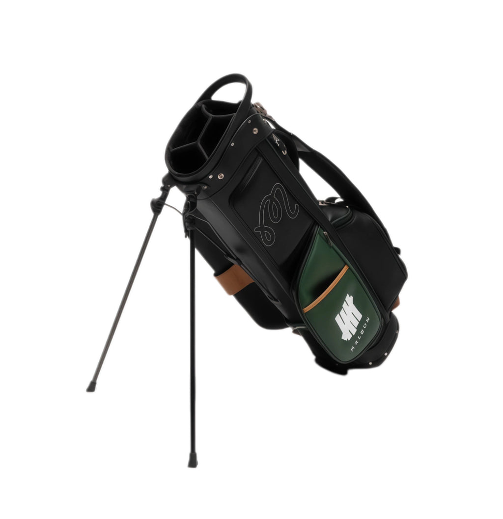 Angled shot of Malbon Golf stand bag with deployed black dual-leg stand system, highlighting lightweight structure, top carry handle, and sleek black/green color blocking design.