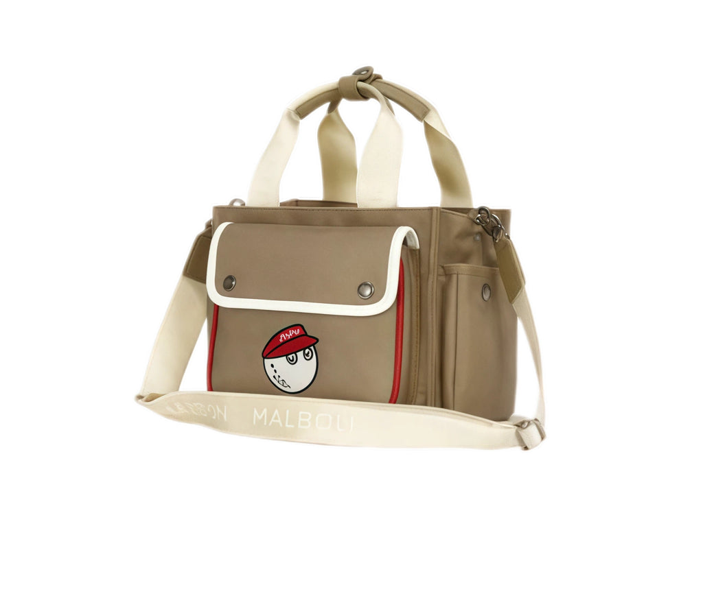 Angled view of beige Malbon golf tote bag with red-capped cartoon golf ball patch, adjustable cream shoulder strap labeled "MALBON", and structured sides.